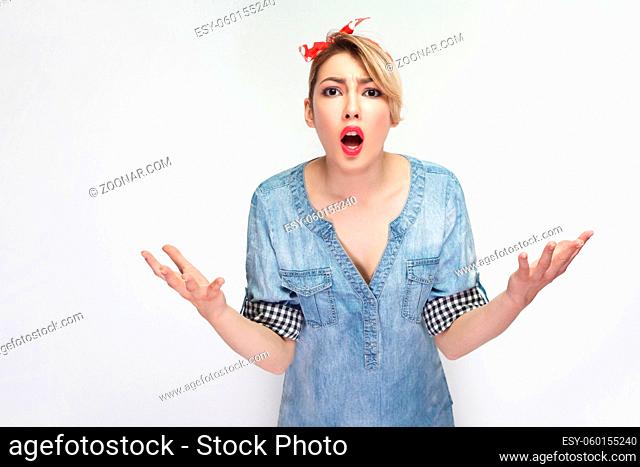 Portrait of shocked angry beautiful young woman in casual blue denim shirt standing with raised arms and looking at camera and asking