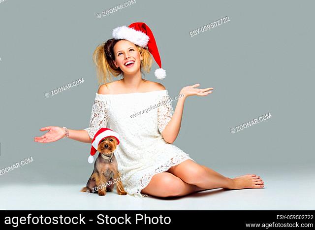 Beautiful happy girl in lace dress and red santa cap sitting with pretty yourkshire terrier in christmas hat. Studio shot over grey background