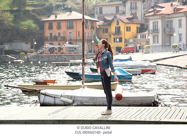 Young woman standing on pier eating ice cream cone at lake Mergozzo, Verbania, Piemonte, Italy