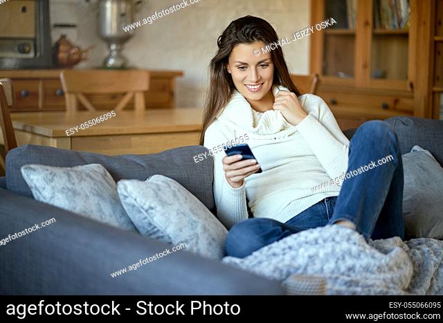 beautiful young smiling woman in white sweater texting on her phone on a gray sofa