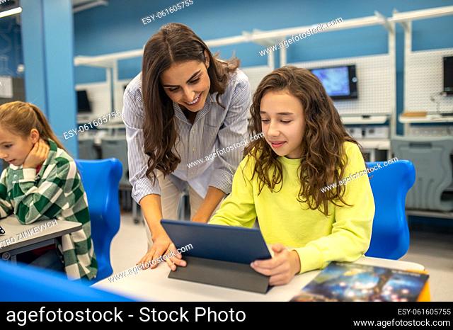 Good result. Smiling teacher leaning approvingly looking at tablet of girl sitting studying at desk during lesson