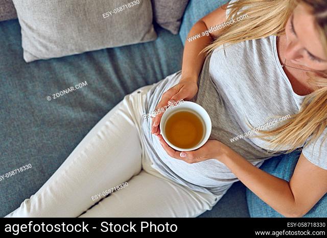 Top view of a young pregnant woman reclining on a comfortable sofa at home with a large mug of hot tea in a close up on her belly