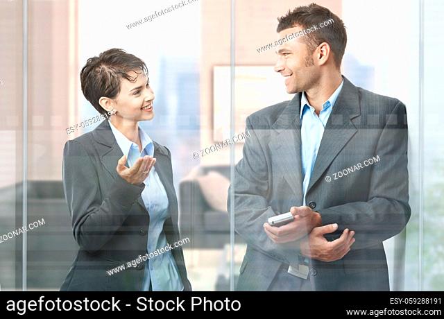 Two happy businesspeople standing in modern office with glass walls, looking at each other, smiling