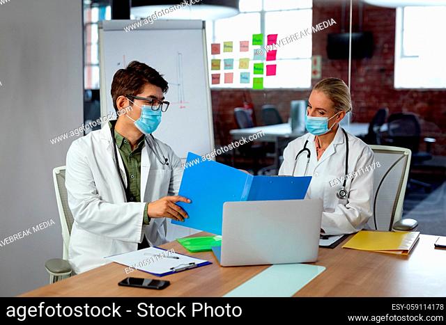 Diverse male and female doctor wearing face masks sitting in hospital office discussing paperwork