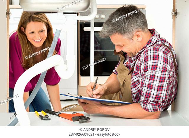 Happy Mature Woman Looking At Male Plumber Writing On Clipboard