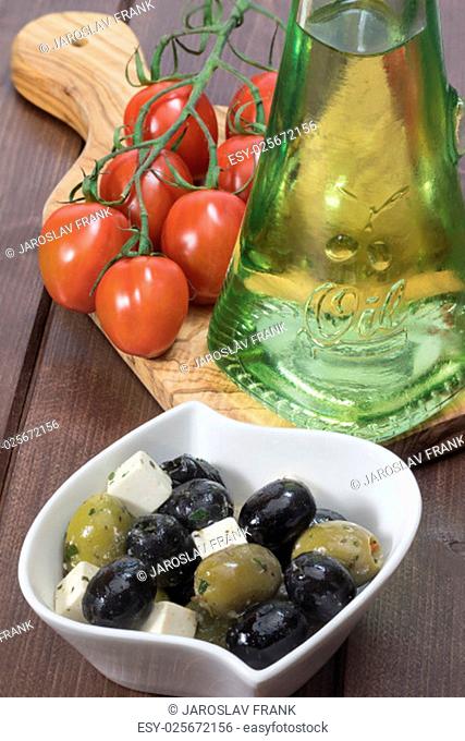 White bowl with marinated cheese, black and green olives is placed on a wooden desk. Tomatoes and bottle of oil are lying on a desk of olive wood