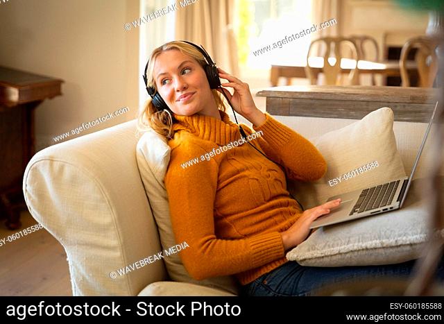 Smiling caucasian woman relaxing on couch in living room wearing headphones and using laptop