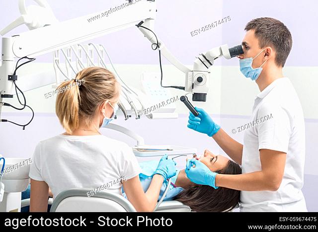 Pretty girl with patient bib on a dental chair and dentist with assistant who sit next to her. Man looks on her teeth using dental microscope and holds a dental...