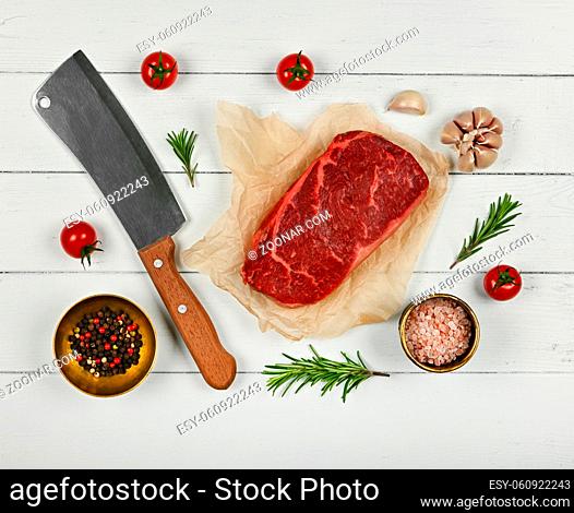 Close up one aged prime marbled raw sirloin beef steak on brown paper parchment wrapping, with cleaver knife and spices, over white wooden table background