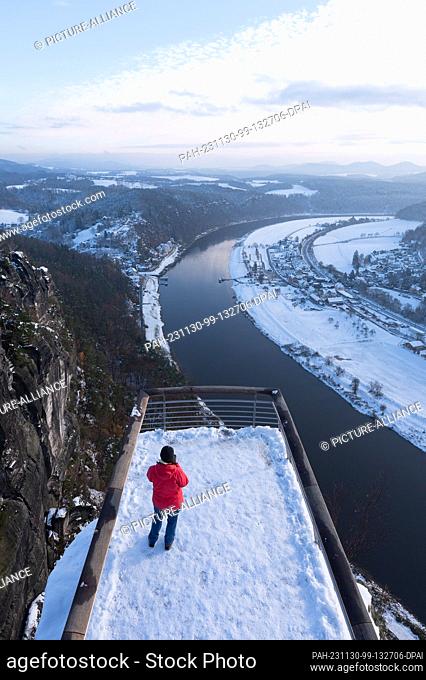 30 November 2023, Saxony, Lohmen: A passer-by stands on a viewing platform on the Bastei rock in the snow-covered Saxon Switzerland National Park
