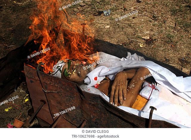 Flames from high pressure paraffin burner around the face of mans body in white shroud laid out on metal grating in simple village Hindu cremation ceremony