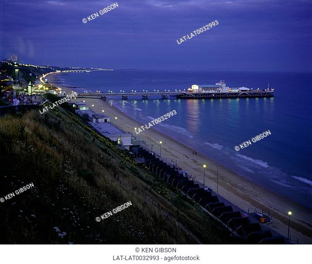 Seaside town. Coast. Coastline. Long beach. Curved bay. Pier buildings lit up at night. Flat calm sea. Reflectionso f lights. Buildings. Headland
