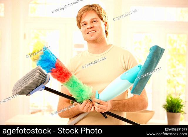 Young man smiling confidently with cleaning tools, holding mop, broom and flannel