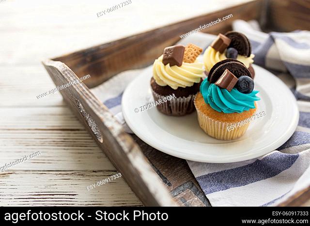 Tasty multicolored muffins and cupcakes on the white plate on a wooden tray with towel. Decorated with different candies