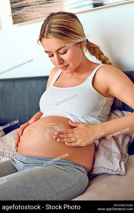 Young pregnant woman rubbing moisturising cream on her belly to moisturise her skin and reduce the possibility of stretch marks after childbirth