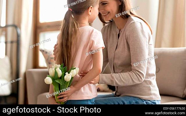Cute little daughter hold hiding flower bouquet behind back making birthday surprise for smiling mom, young happy mother and preschooler girl kid hug having...