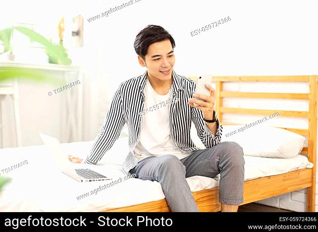 Handsome young man in casual clothes sitting on the bed, with a laptop in bedroom, play mobile games and chat on the phone