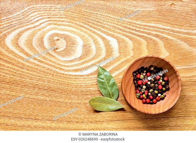Fragrant colorful pepper in a wooden bowl and two dried bay leaves on a light wooden table. Top view