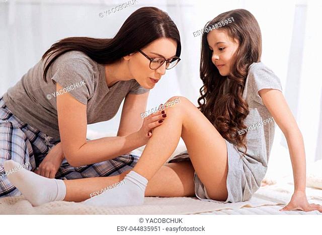Receiving emergency help. Skillful optimistic young woman sitting at home while helping little sister and putting adhesive bandage on her knee