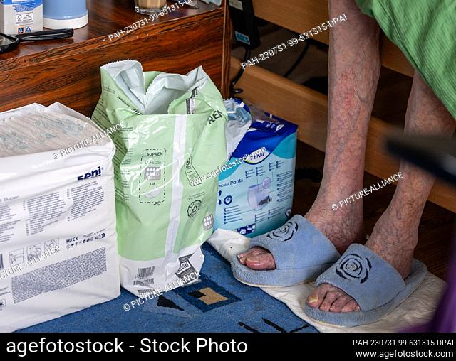 PRODUCTION - 31 July 2023, Berlin: A senior in need of care sits on the bed next to diapers and incontinence pads during a caregiver's visit to her home