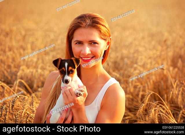 Young woman smiling, holding Jack Russell terrier puppy, sunset lit wheat field in background