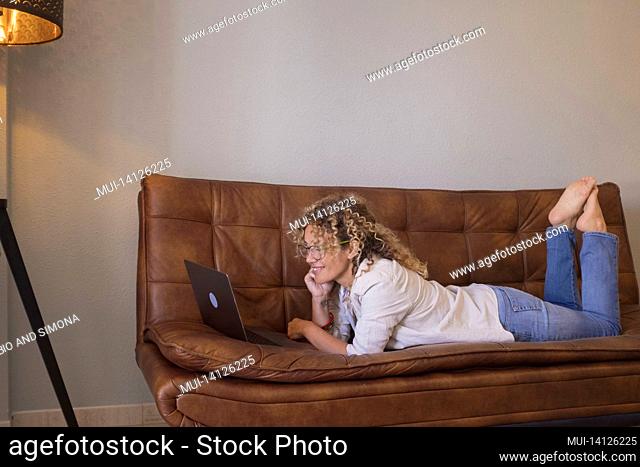 profile of smiling woman in eyeglasses and curly hair watching media content using laptop on comfortable leather sofa at home