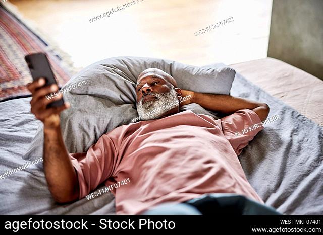 Bald man with hand behind head using smart phone lying on bed at home