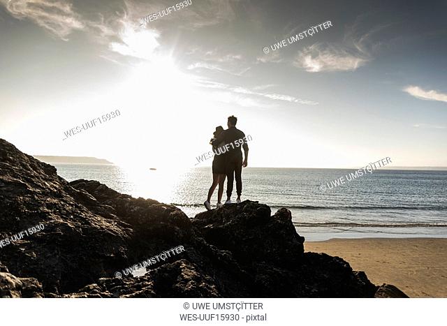 France, Brittany, rear view of young couple standing on rock at the beach at sunset