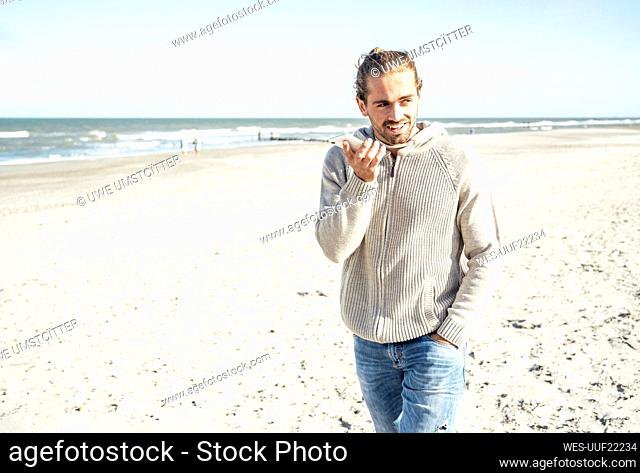 Man with hand in pocket talking on mobile phone while walking at beach during sunny day