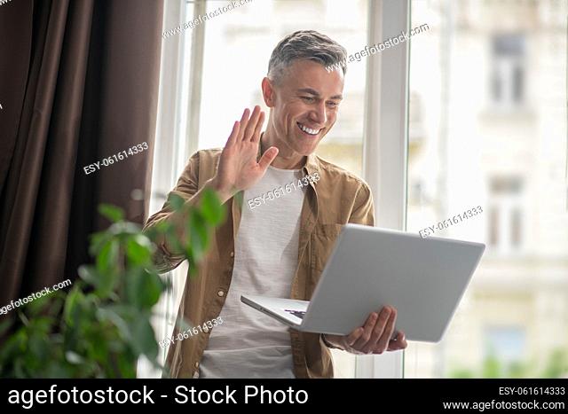 Video call. Joyful man standing in room holding laptop greeting waving his hand smiling at screen