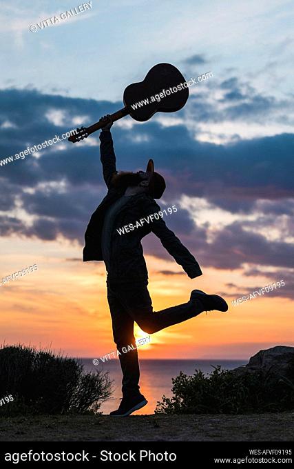 Young man with hand raised holding guitar during sunset