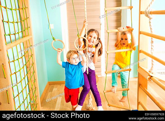 Three active children in colorful t-shirts, excercising together, hanging and climbing on a rope, ladder and rings at their come sport complex