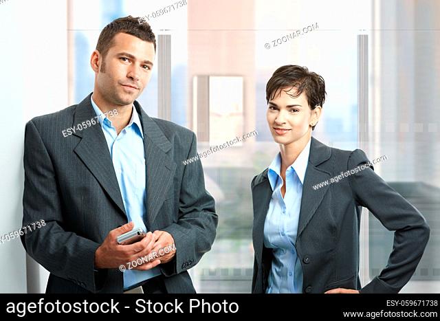 Two young businesspeople standing in modern office with glass walls, looking at camera, smiling