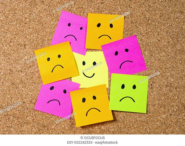 smiley cartoon face expression on yellow post it note surrounded by sad and depressed faces on cork message board in happiness versus depression and smile...