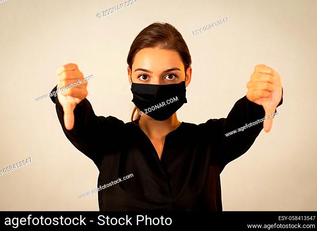 Unhappy young woman wearing black cloth face mask expressing her negative emotions. Girl in dark shirt with covered mouth and nose showing thumbs down