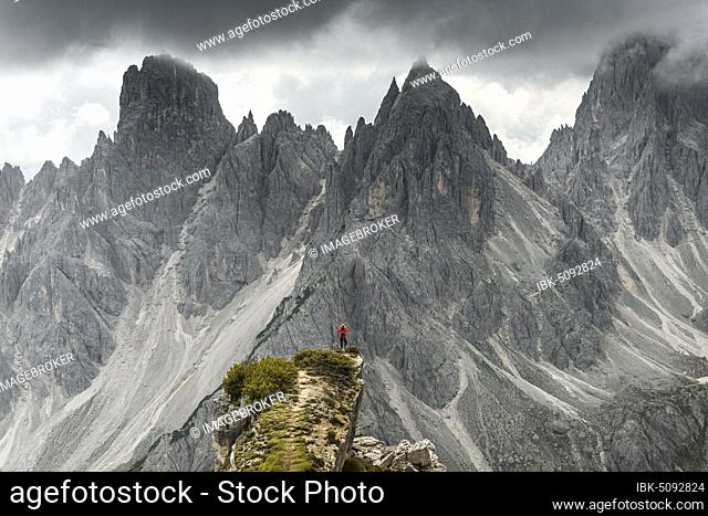 Man with red jacket standing on one degree, behind him mountain peaks and pointed rocks, dramatic clouds, Cimon the Croda Liscia and Cadini group