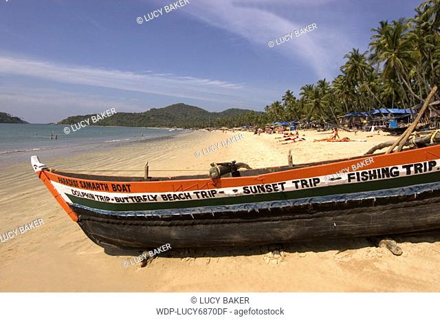 Fishing boat in foreground.Pale golden swathe of Palolem Beach in background. People on beach and in sea. lined in palm trees
