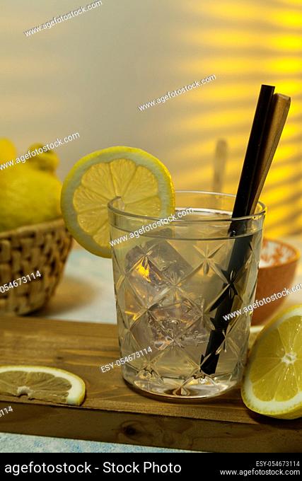 lemonade glass with ice, spoon and straw on cutting board, cut lemons and basket with lemons on background with sunlight entering through rear window