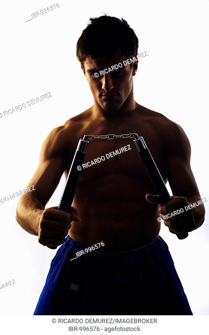 Young fighter with bare upper body holding nunchaku, backlight
