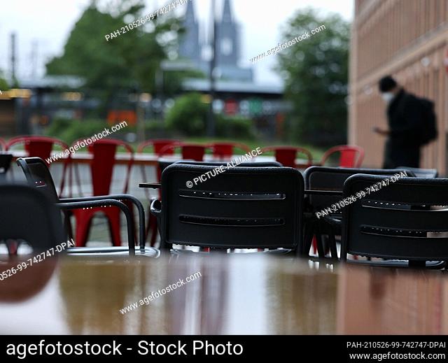 26 May 2021, North Rhine-Westphalia, Cologne: Chairs and tables stand outside a restaurant in the rain, with the cathedral in the background