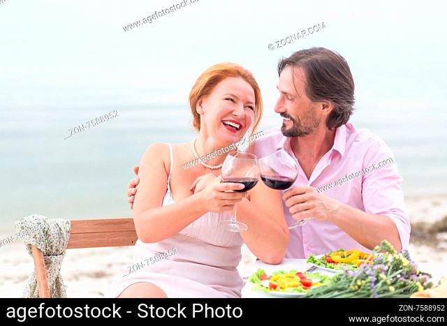 Happy mature couple outdoors near by sea. Red-haired woman laughing with her boy-friend, they both are drinking red wine
