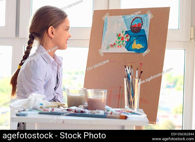 The girl happily looks at her finished drawing hanging on an easel