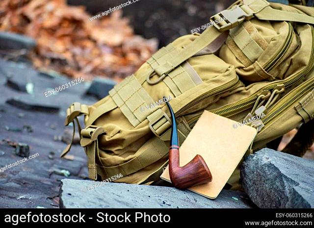 Close up product photography style shot of a green army tactical backpack, a travel notebook and a pipe disposed on rock steps in the woodlands