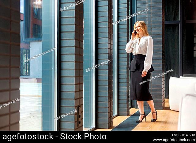 Full length portrait of beautiful elegant businesslady with blond hair in white shirt and skirt looking out window of business center and talking on cellphone...