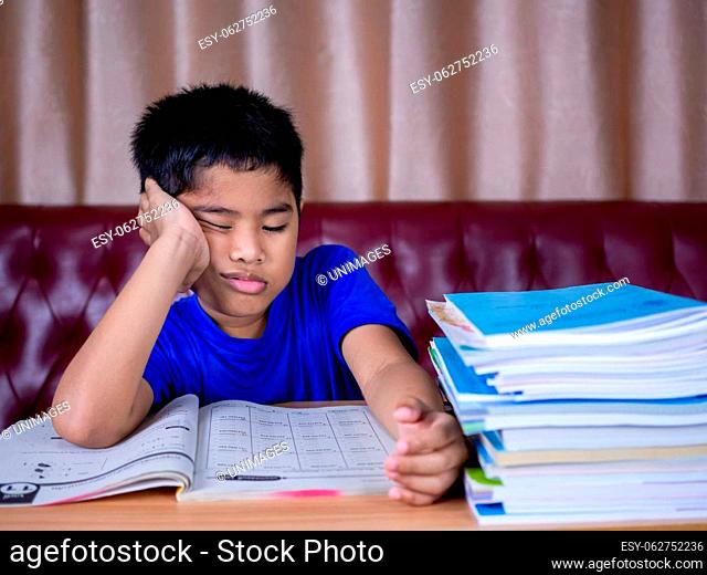 boy doing homework and reading on a wooden table with a pile of books beside The background is a red sofa and cream curtains