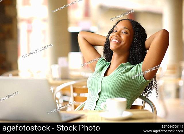 Relaxed happy black woman with laptop resting sitting in a bar terrace looking at you