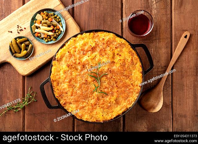 Homemade shepherd's pie in a cooking pan with pickles and wine, shot from the top on a dark rustic wooden background with herbs