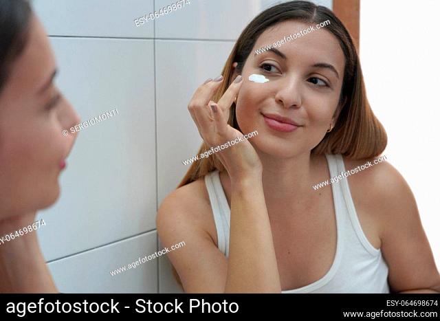 Skin care routine at home. Portrait of beautiful girl applying cream under eye with finger. Dark circles and anti-wrinkle concept