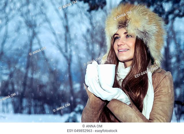 Portrait of a beautiful young girl wears a stylish hat standing in a snowy park and warming hands on a hot mug of tea, enjoying the beautiful winter view