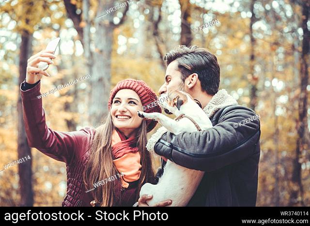 Woman and man with their dog on autumn walk taking a phone selfie posting it online on social media